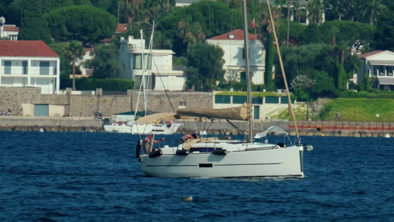 Barca a vela che naviga in acque blu vicino a ville costiere e yacht ormeggiati, catturando la serenità di una vacanza estiva, Antibes, Francia