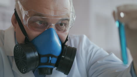 a scientist at a pharmaceutical company developing drugs wearing a blue respirator and safety glasses holds a new medicine a glass tube with a blue liquid in his hands and looks at it. high quality 4k footage