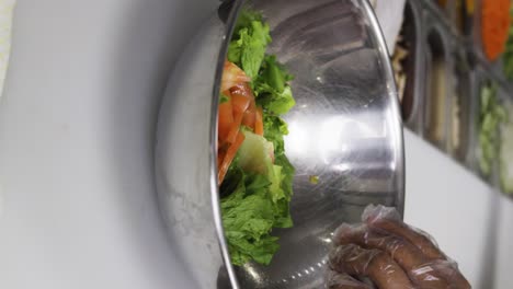 vídeo de cerca vertical de un cocinero con guantes de plástico haciendo una ensalada en un cuenco metálico