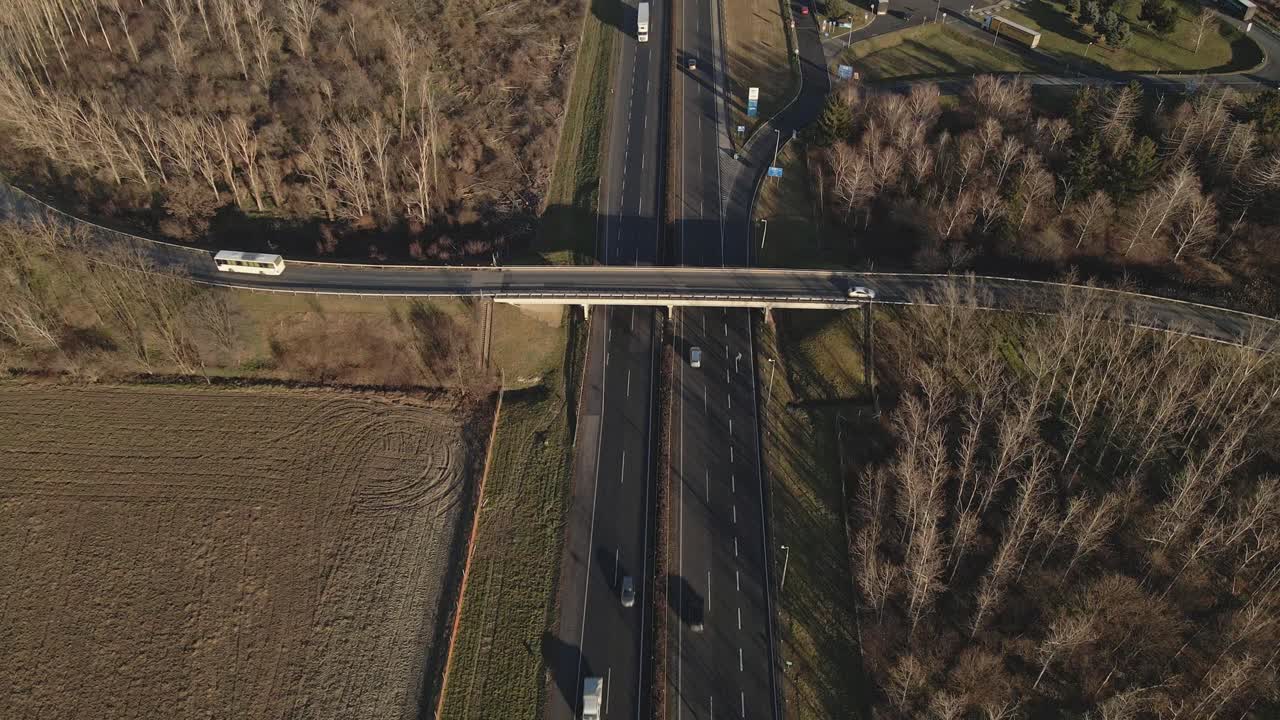 An aerial view of a highway overpass shows vehicles traveling on both the main road and the bridge above. The surrounding landscape features fields, trees, and a nearby intersection.
