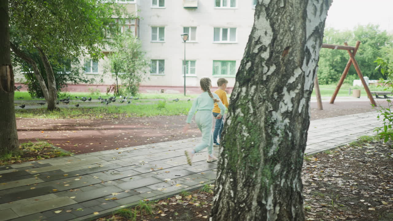 Girl adjusts shoe while hurrying to catch up with brother on wet park pavement, surrounded by leafy trees and distant pigeons, near residential building in soft natural light after rain