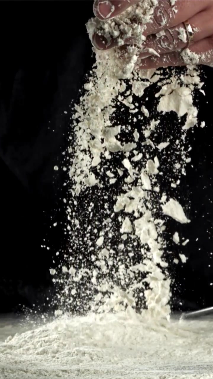 Mano de chef tamizando harina blanca en cámara lenta sobre fondo negro