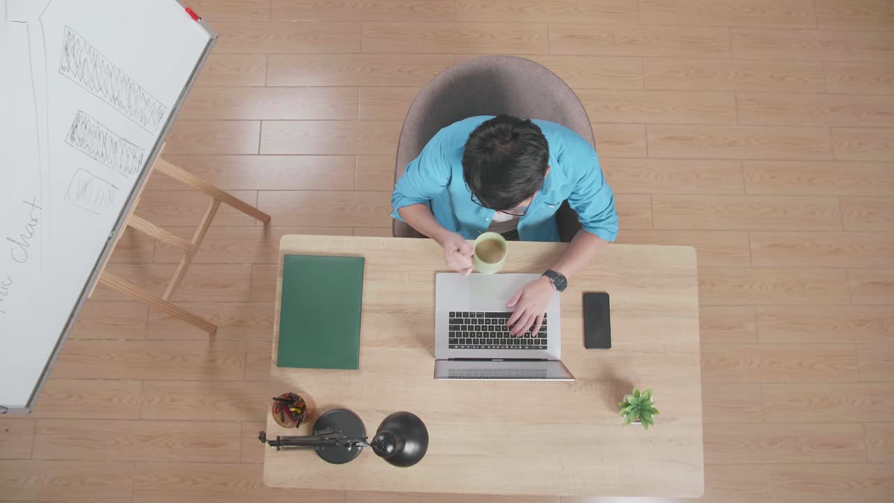 vista superior zoom out toma de un hombre bebiendo café y escribiendo en una computadora portátil al lado del teléfono inteligente mientras está sentado en el escritorio de madera junto a una pizarra en casa
