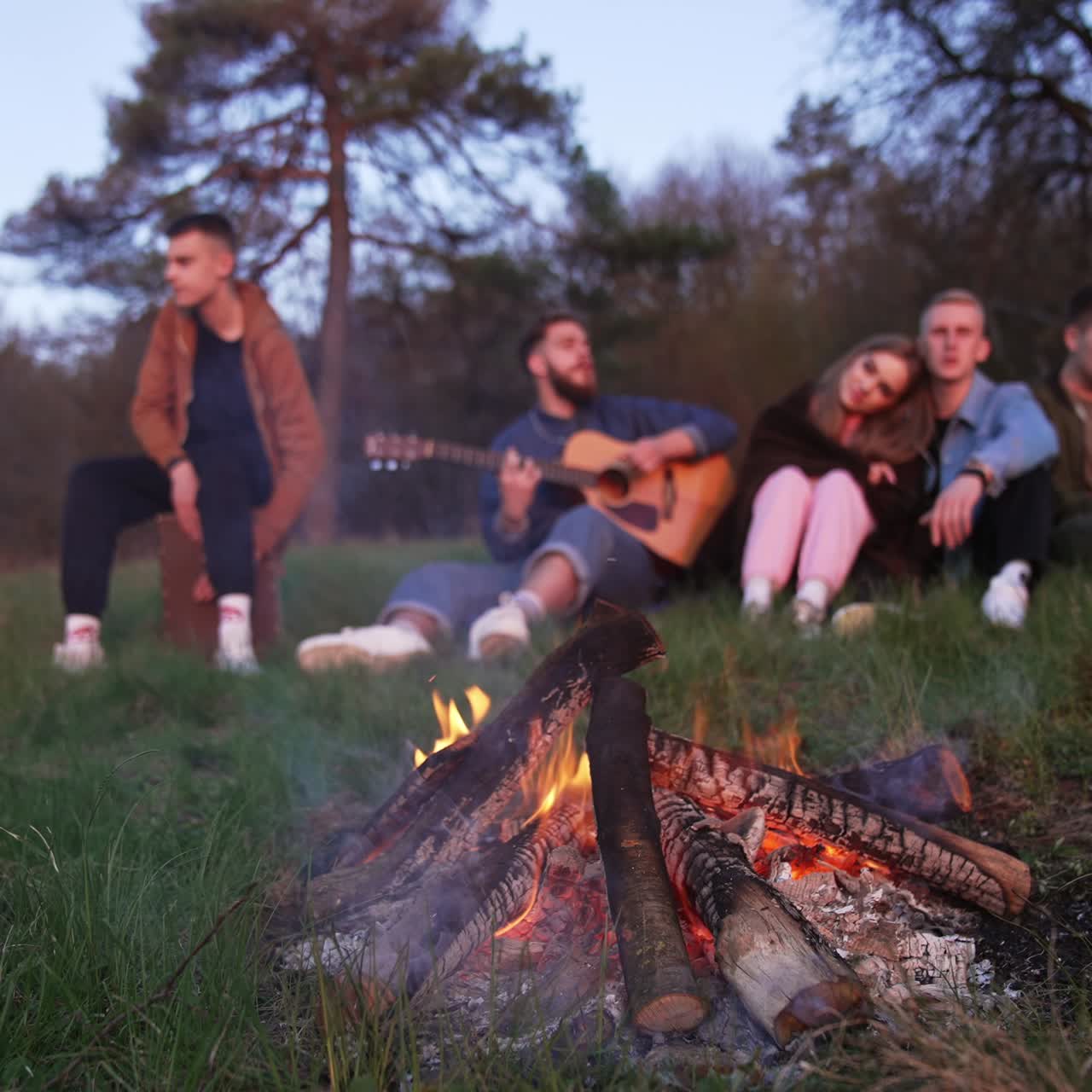 Pieces of wood burning slowly in the campfire. Company of youngsters sitting on the grass, playing guitar and singing. Blurred background