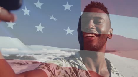 animation of flag of usa over smiling african american man using smartphone on beach