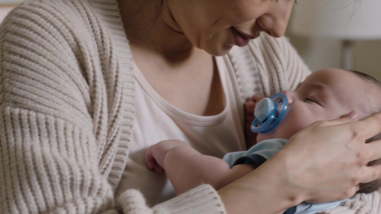 Cradling mother in ribbed beige cardigan soothing drowsy infant on bed in blue onesie, pacifier