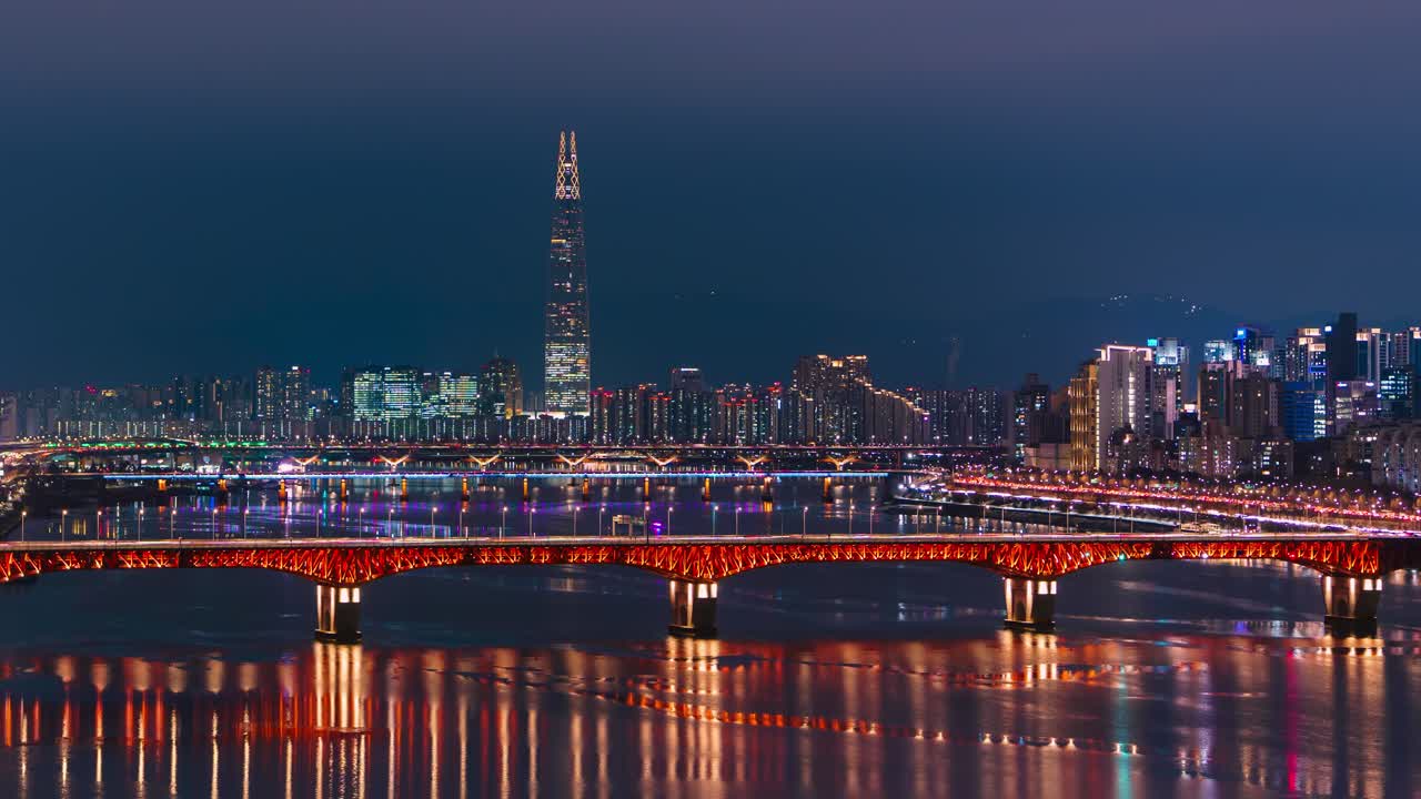 Un timelapse svela il Ponte Seongsu illuminato da luci rosse con l'imponente Lotte World Tower che perfora lo skyline di Seul tra i riflessi del fiume Han e il bagliore circostante della città di grattacieli.