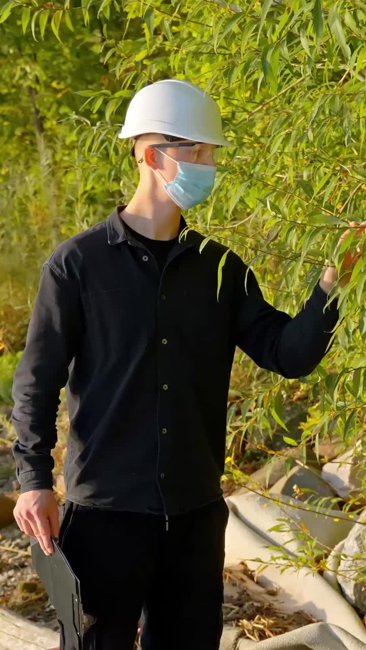 Scientist in dark clothes inspects rubbing leaves at sunset, taking notes on clipboard, vertical