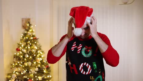 man wearing festive jumper in front of christmas tree thrown santa hat and blows snowball end off his face pointing at camera and smiling