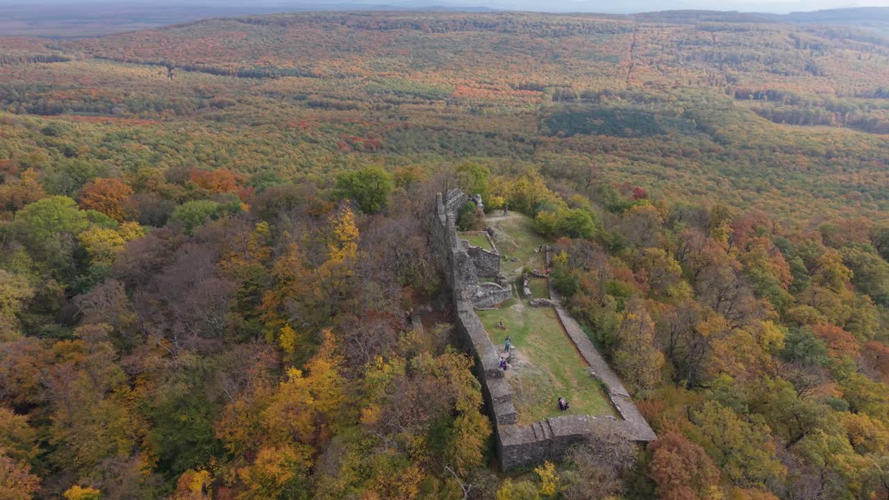 A drone view of historic stone castle ruins perched on a forested hilltop during peak autumn colors. The surrounding landscape is filled with vibrant foliage in shades of orange, yellow, and green