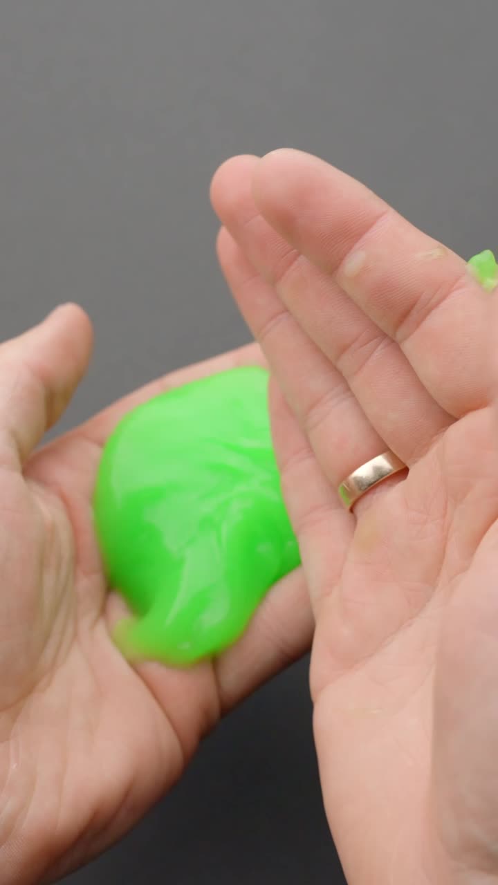 Vertical close-up image of a person squeezing and stretching bright green slime in their hand, used for ASMR sensations and stress relief through tactile play and visual softness