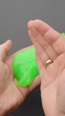 vertical close-up image of a person squeezing and stretching bright green slime in their hand, used for asmr sensations and stress relief through tactile play and visual softness