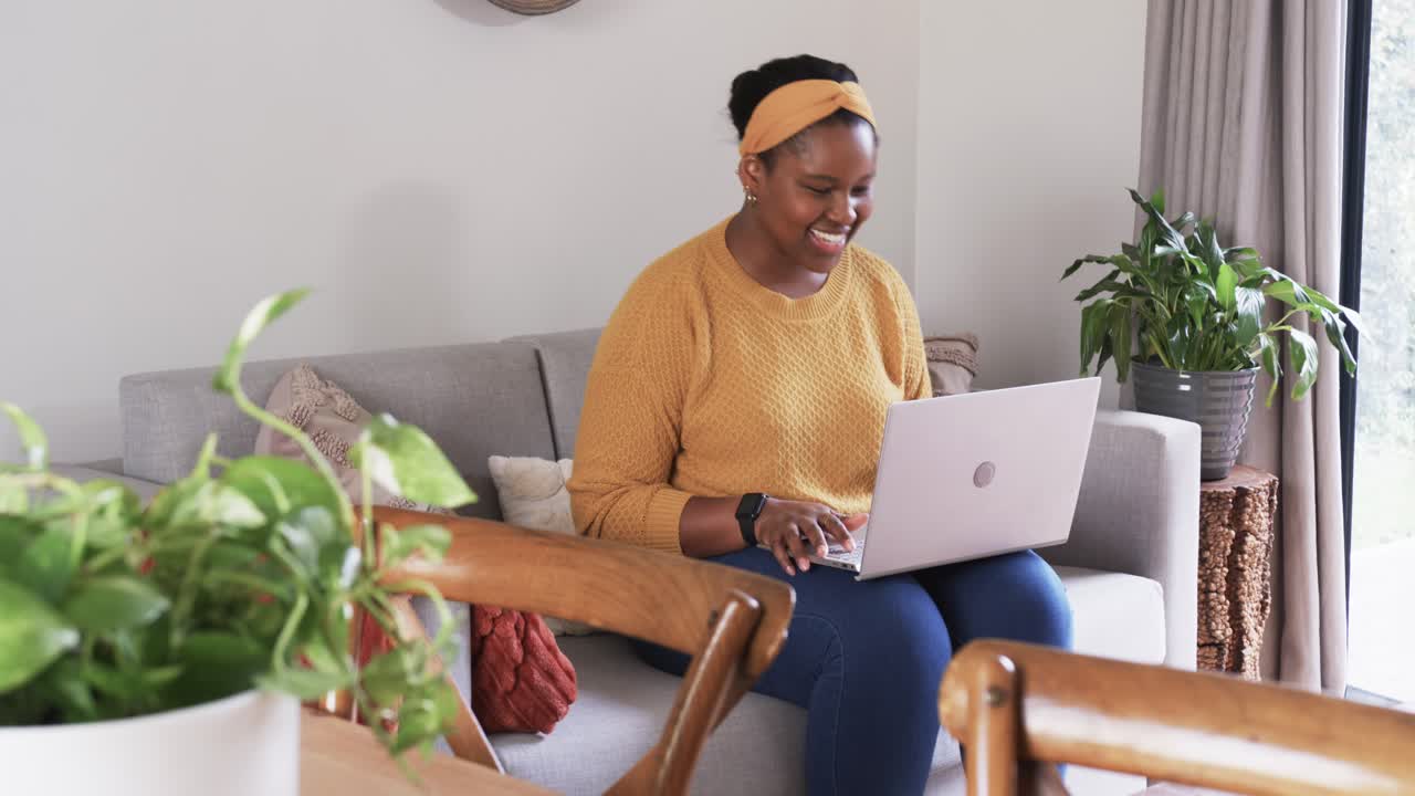 Leaning forward on sofa, African American woman lifting laptop onto lap, typing and smiling at home