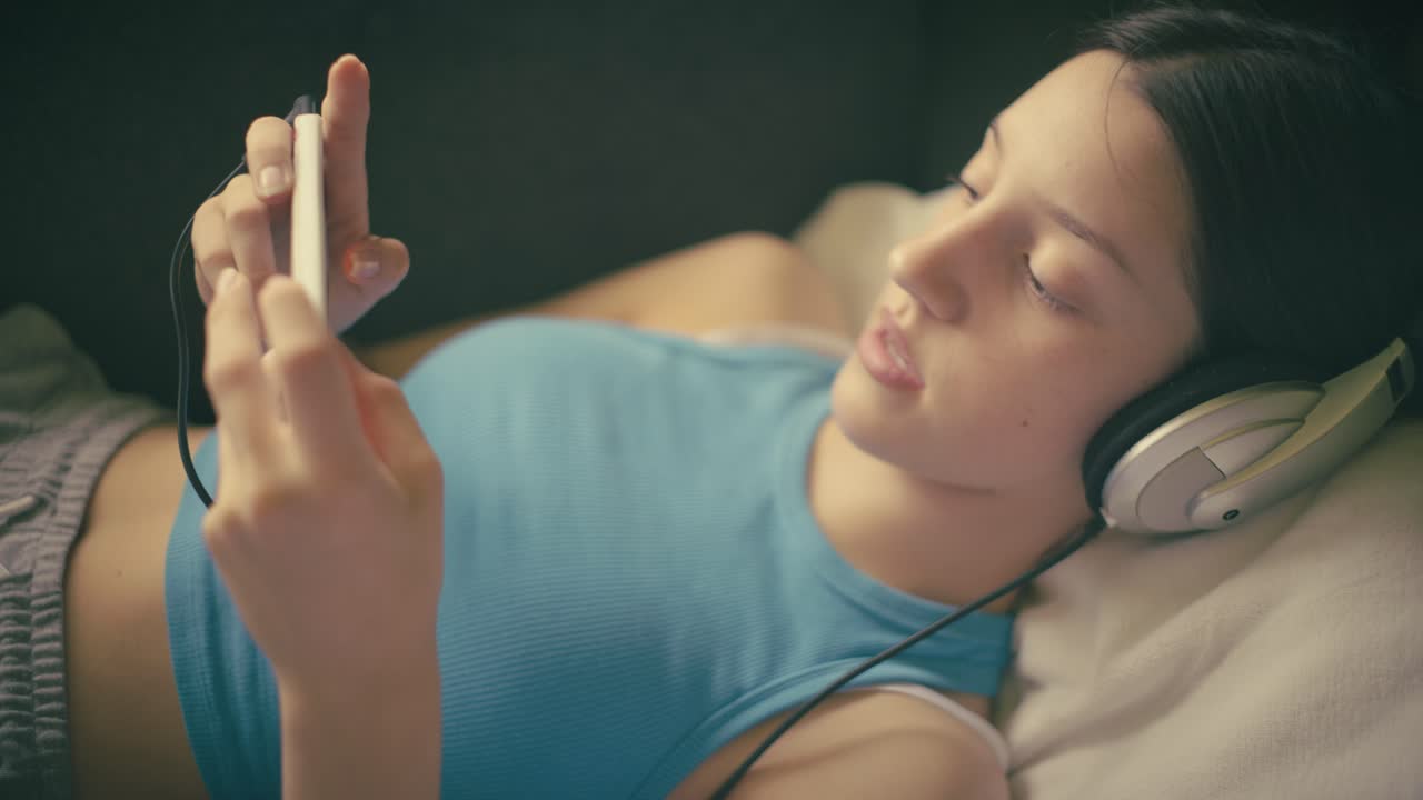 Teen girl relaxing at home, listening to music with headphones and using her smartphone. A calm, intimate moment of digital leisure in a cozy setting.