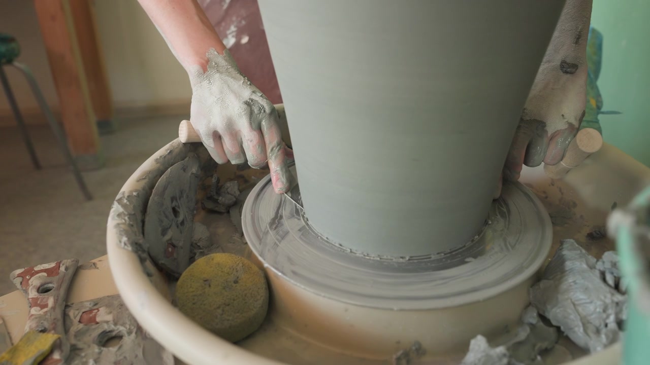 Kaukasische Hände beschneiden nasse Tonbasis auf der Töpferscheibe, mit Schwamm und Eimer in der Nähe. Nahaufnahme von Druck und Rhythmus, präzise Werkzeugbewegung entfernt Überschuss, taktile Studientechnik und konzentriertes Handwerk.