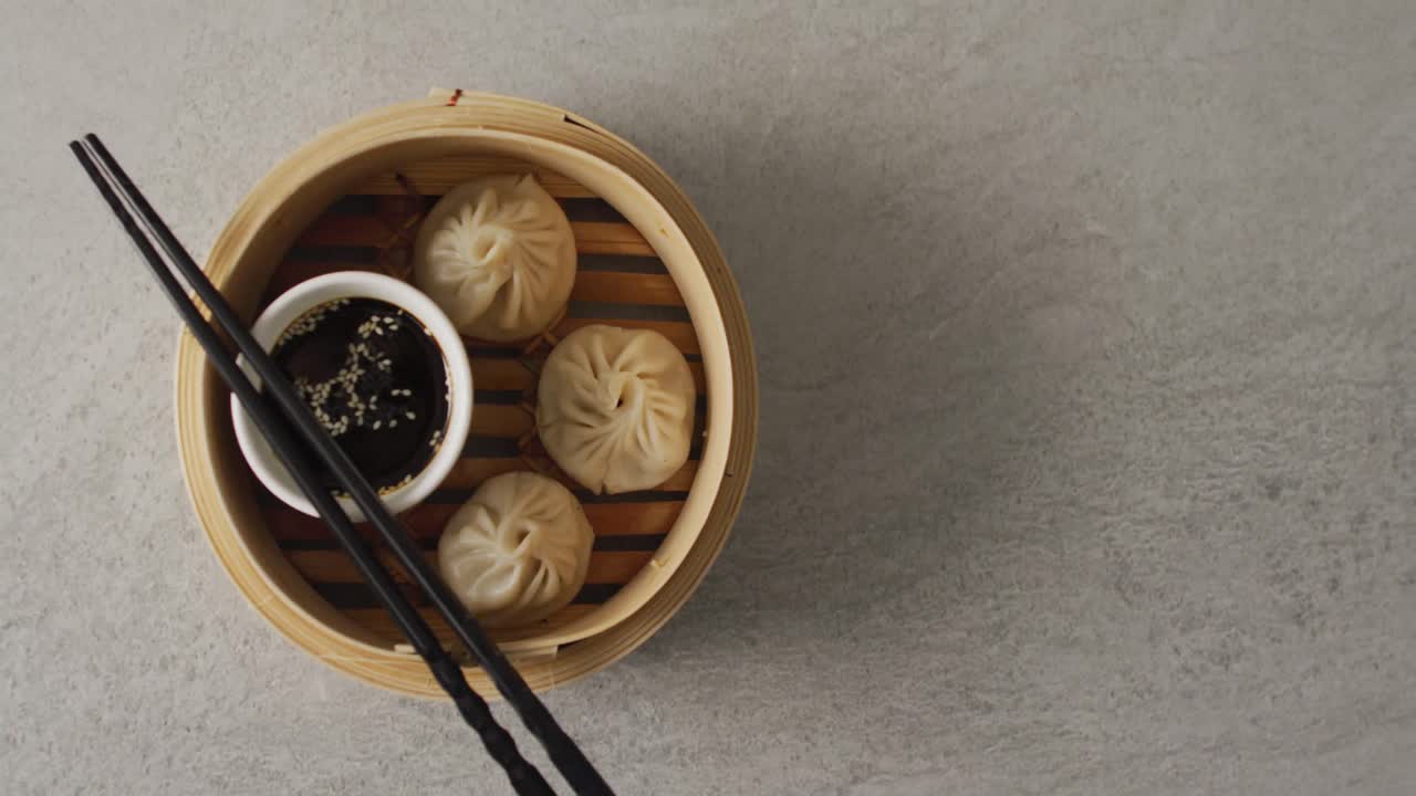 Komposisi kukusan bambu dengan pangsit dim sum dan sumpit dengan kecap asin di latar belakang abu-abu