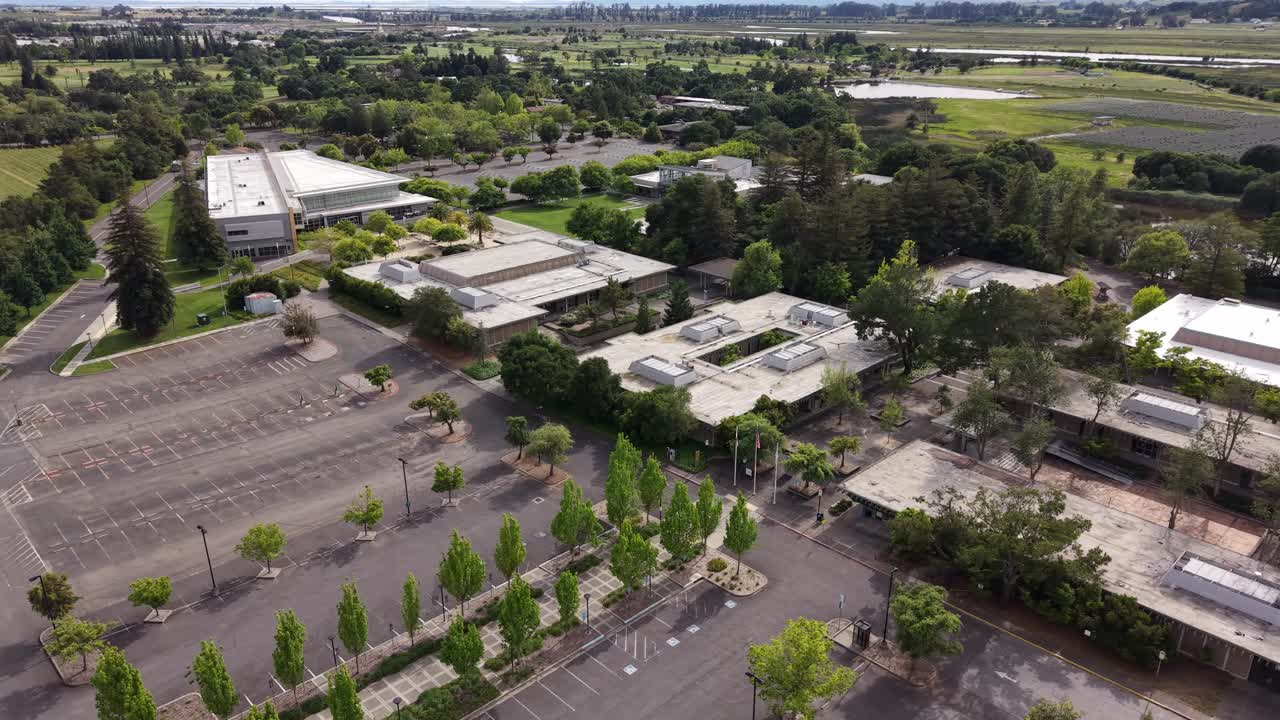 Vista aérea en picado del campus de Napa Valley College en Napa, California, mostrando edificios modernos rodeados de exuberantes árboles verdes y un amplio estacionamiento vacío en un día soleado.