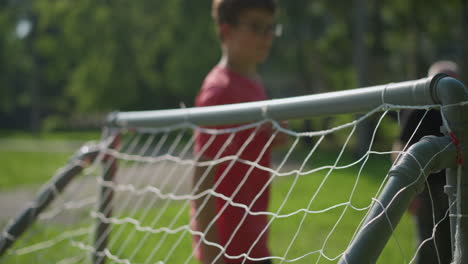 a ball is shot against a goal post as a young goalkeeper in red moves to catch it, the grandfather watches from the background