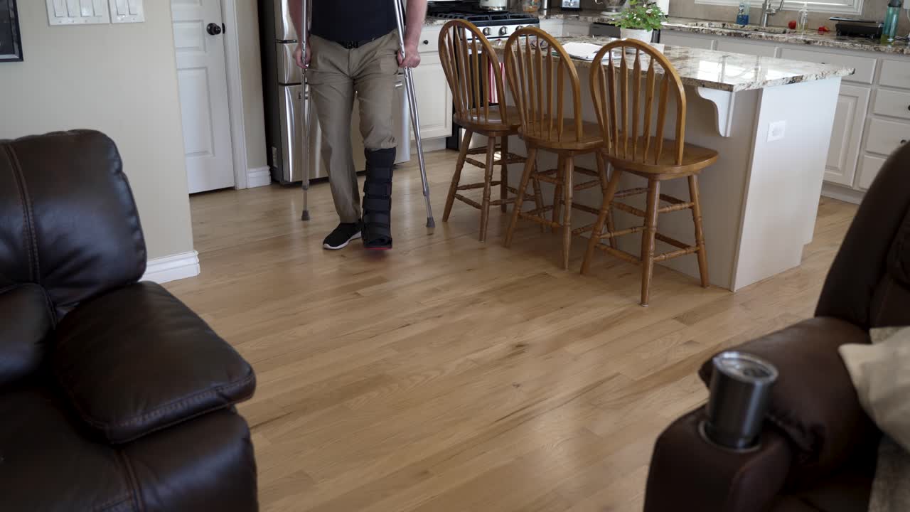 hombre con una pierna rota en una bota ortopédica usa muletas para moverse por su casa - inclinado hacia abajo