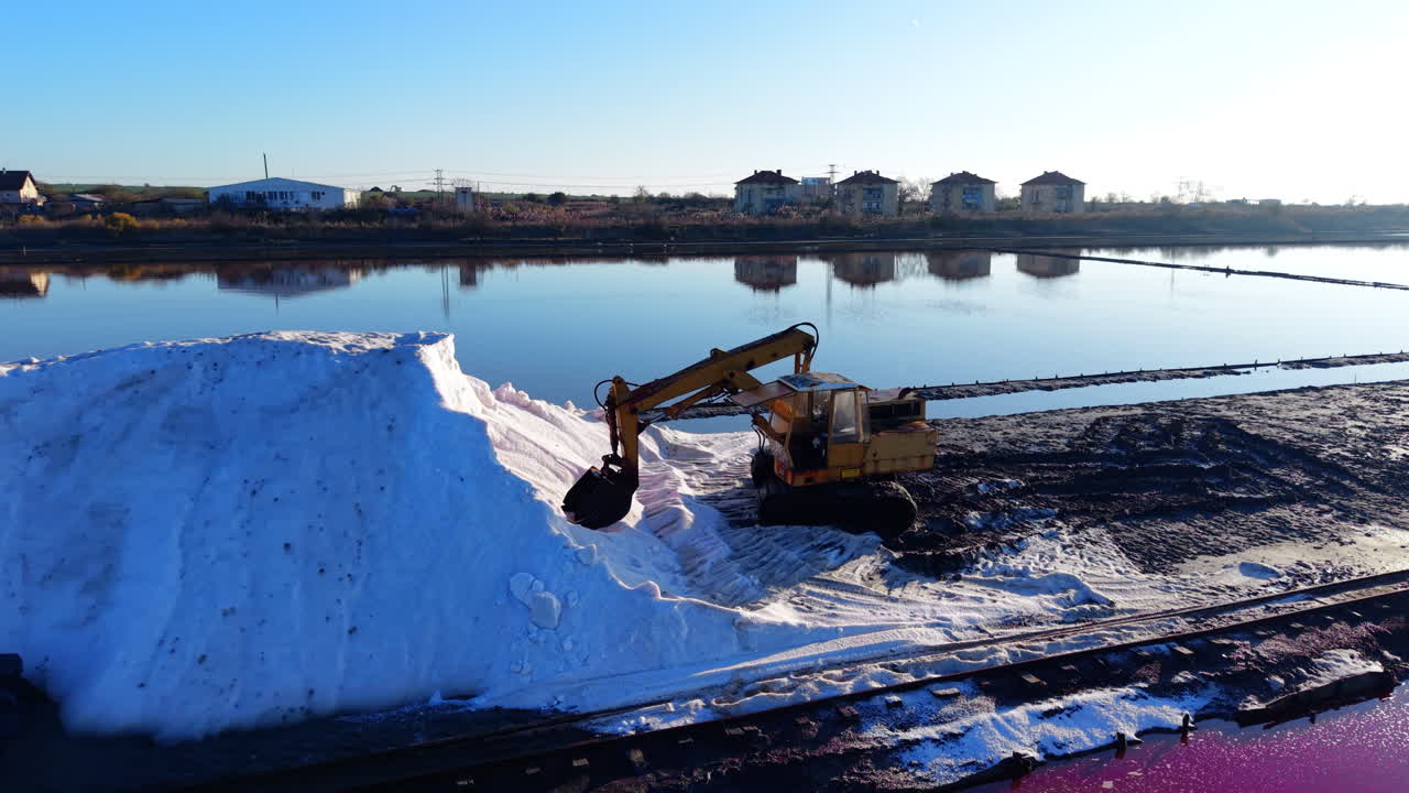Macchinari industriali al lavoro su un'enorme montagnola di sale bianco. Le acque calme della laguna riflettono il lontano skyline urbano sotto un cielo terso. Escavatore giallo che accumula sale marino