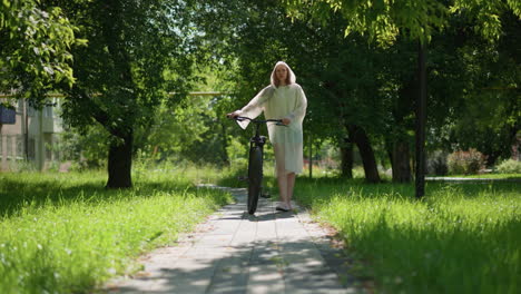 woman in translucent raincoat strolls along sunny pathway with bicycle, surrounded by lush greenery and trees, with building and another person walking in background