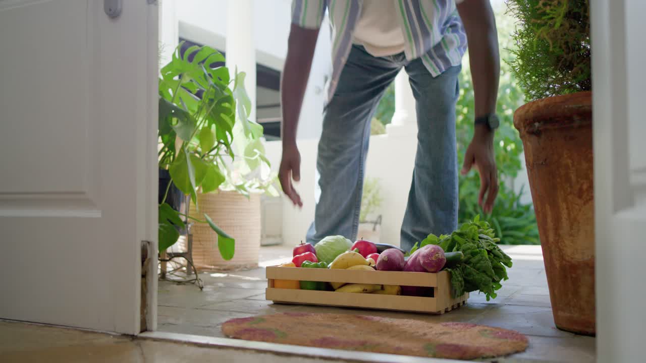 Mature African American male spotting crate on doormat, bending and carrying produce into house