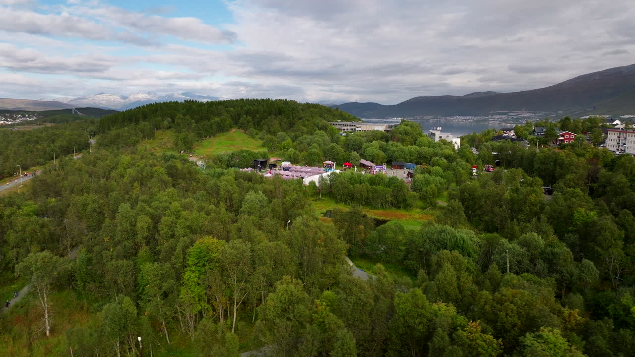 Relay for Life charity event in Tromsø, people walking on track, tents, and stage, with city and mountains in background, Norway