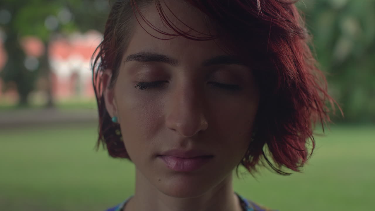 High-quality 4K footage of a serene young woman with vibrant red hair and closed eyes. Shot outdoors in soft, natural light. Ideal for mindfulness, beauty, and alternative lifestyle advertising