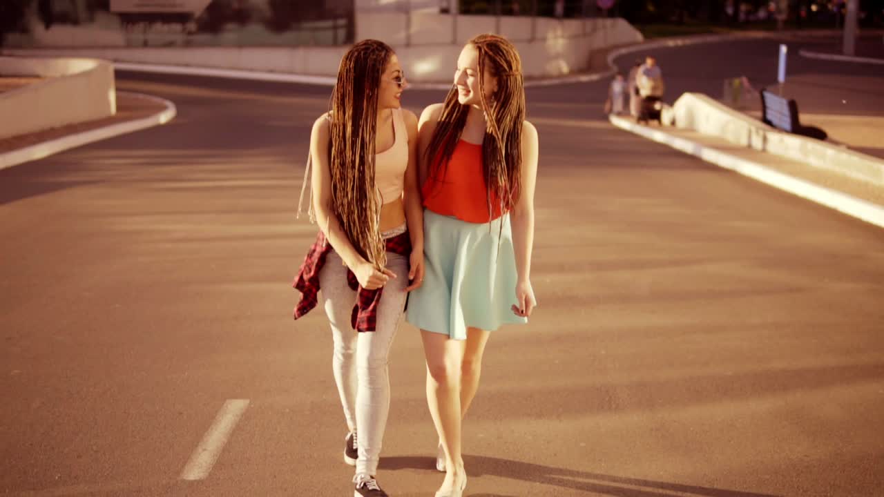 Two happy women with dreads walking on the empty road and talking in summer. Two hipster girls laughing and hugging each other