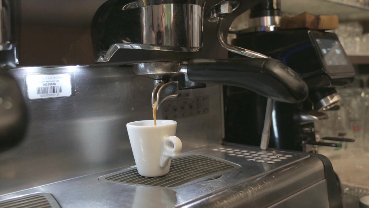 Estrazione di un caffè espresso da una macchina da caffè professionale in un bar. Il caffè scorre bene con una buona crema. Sullo sfondo si intravede un macinacaffè e altri accessori da bar