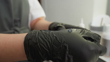 beautician wearing black gloves squeezes liquid substance from bottle onto glove, preparing for application with nearby brush in clean, organized salon setting with cosmetic tools in background