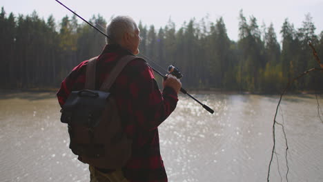 traveller with backpack and fishing rod is standing on high coast of river and admiring nature