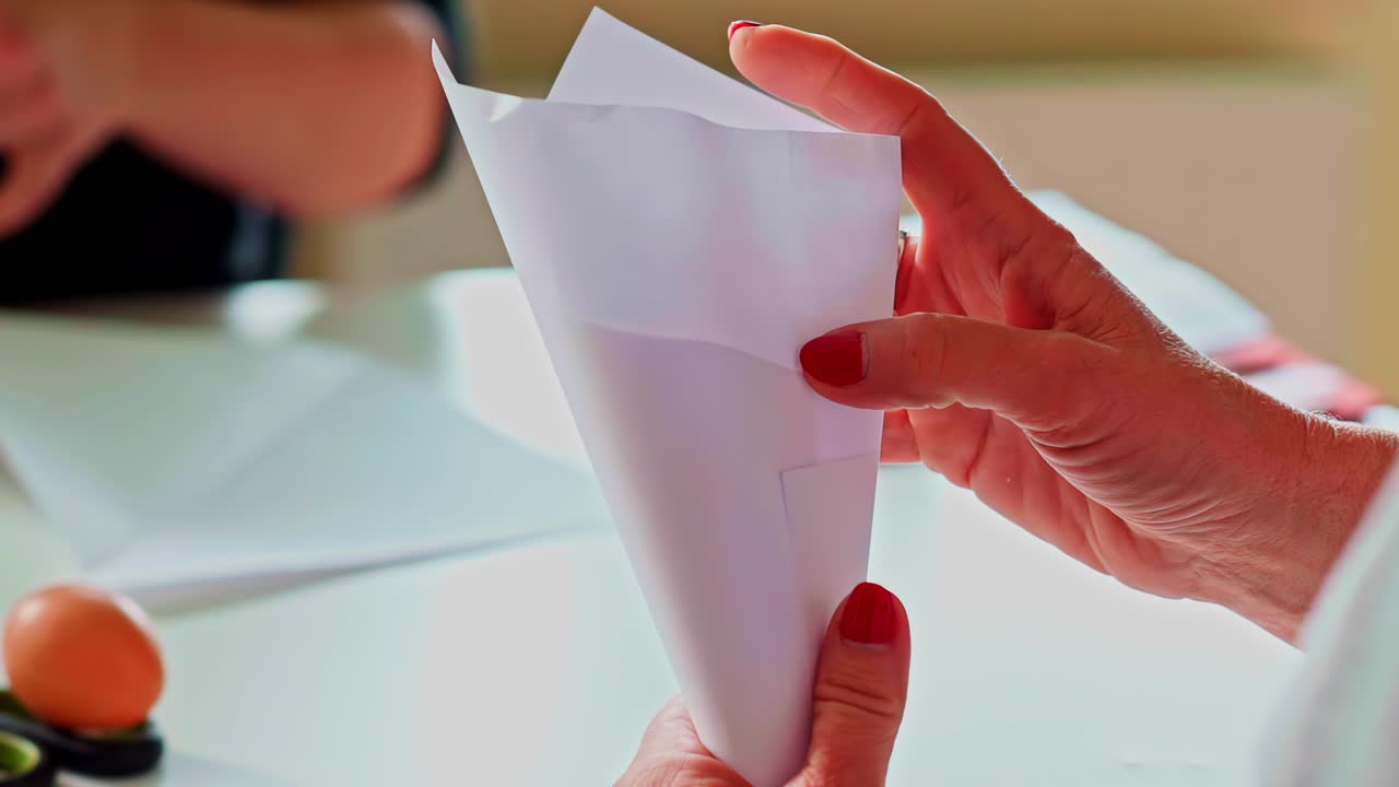 Panoramica lenta verso destra di mani che piegano carta bianca a forma di cono per un progetto artigianale fai da te su una scrivania, processo di lavorazione della carta con forbici e strumenti, tutorial di arti e mestieri fatti a mano