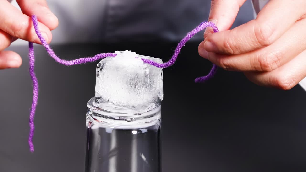 Hands demonstrate lifting an ice cube using a purple string on a glass surface.