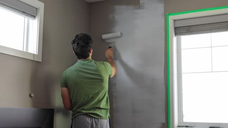 a caucasian man walks into frame with a paint roller covered in white paint and begins to paint the wall beside a window