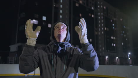 footballer wearing gloves tosses a soccer ball into the air and catches it back during a nighttime practice session on an urban field, bright city lights in the background