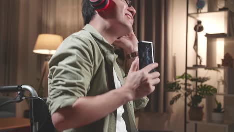 asian man sitting in a wheelchair use mobile phone and listening to music with headphones and dancing in living room