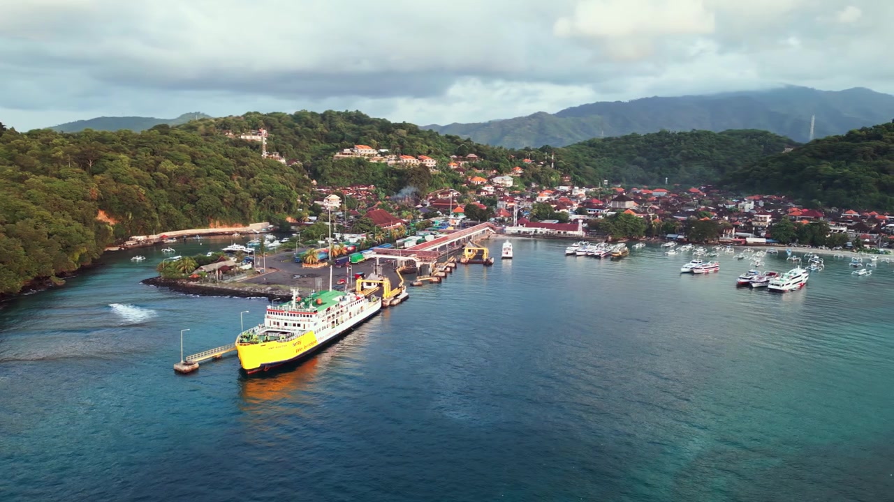 Traghetto ormeggiato a un molo terminale di un porto tropicale accanto a una costa lussureggiante di foresta verde con acqua oceanica di un blu profondo sotto la luce del sole mattutino, ripresa aerea con drone.