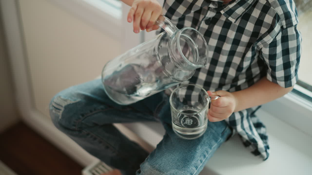 Top down view of young kid in plaid shirt seated by window carefully pouring clear water from glass jar into cup while lifting cup up to drink in bright
