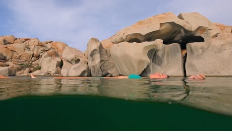 Half-underwater-scene-of-woman-in-blue-bikini-relaxing-floating-on-sea-water-of-Cala-Della-Chiesa-lagoon-with-eroded-granitic-rocks-in-background-of-Lavezzi-island-in-Corsica,-France