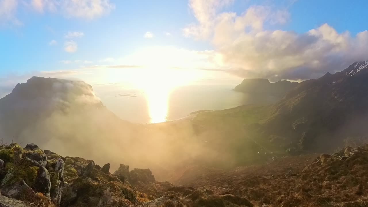 Soft ocean fog rises slowly up mountain slope lit by golden midnight sun over Utakleiv beach