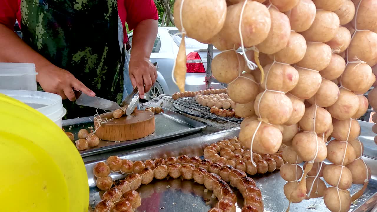 Myyjä valmistaa taitavasti grillattuja makkaroita vilkkaalla katukojulla Kanchanaburissa, Thaimaassa. Kirkas päivänvalo korostaa eloisaa näkymää
