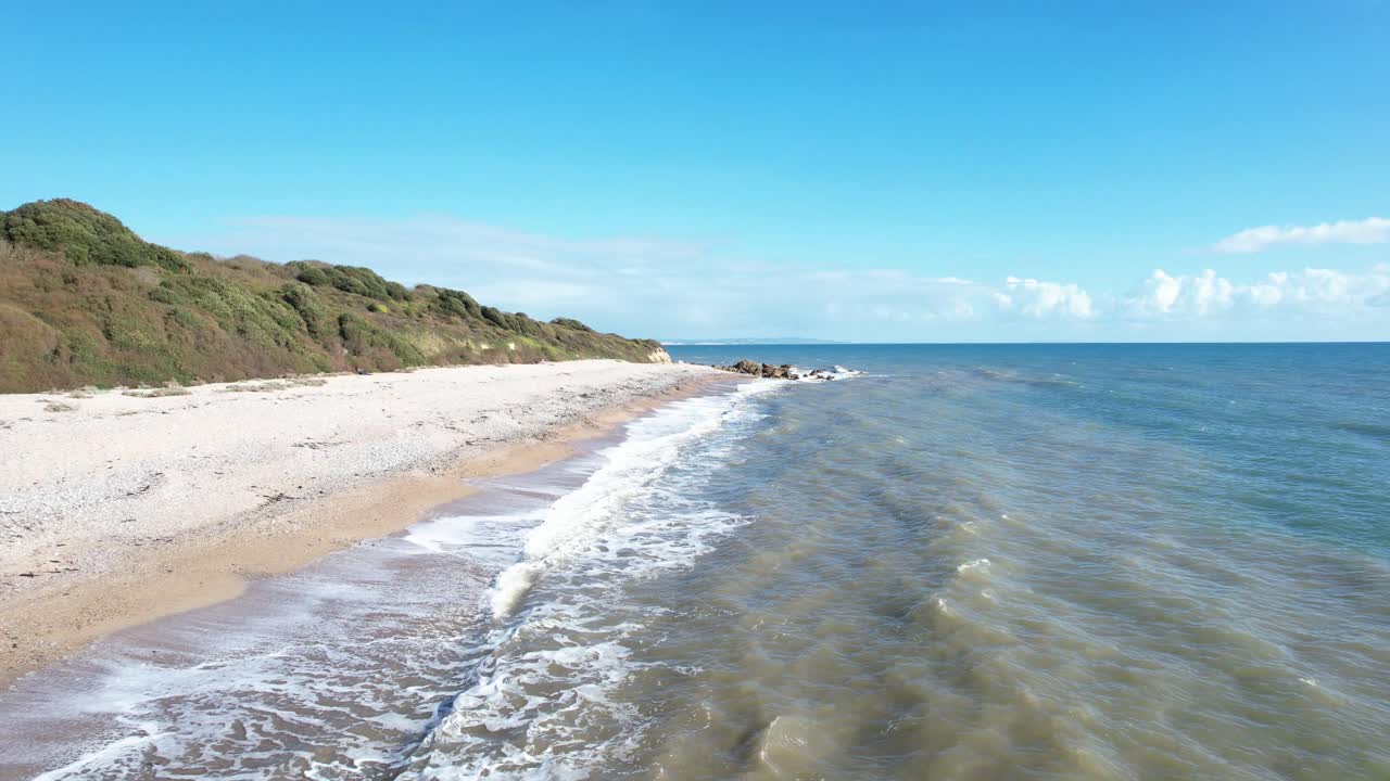 Aerial view of Lyme Regis coastline with gentle waves lapping against the pebbled beach under a clear blue sky. pull back shot