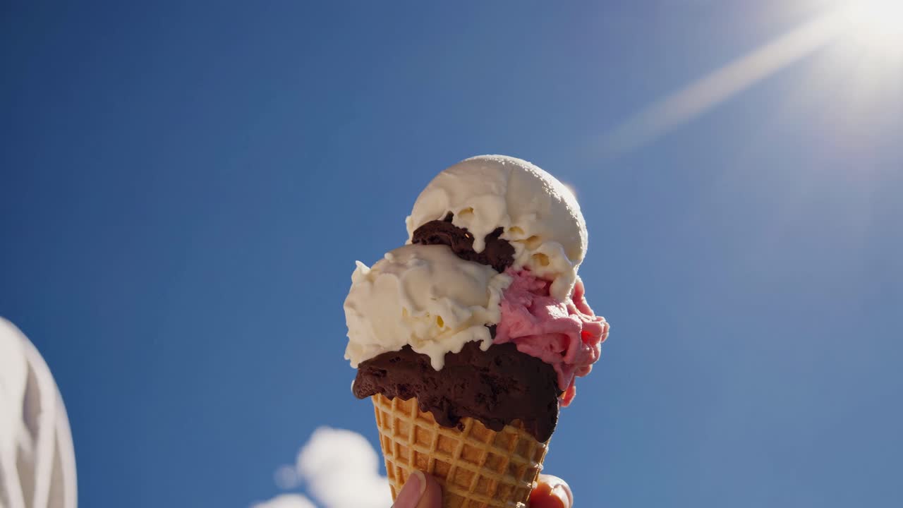 Close-up video angle of a hand holding a colorful ice cream cone against a clear blue sky