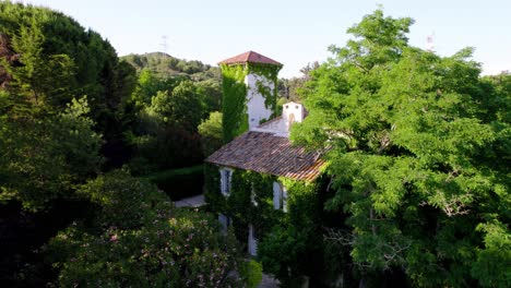 aerial-shot-with-drone-of-a-large-house-hidden-under-foliage,-surrounded-by-trees-on-a-sunny-day,-light-roof-tiles