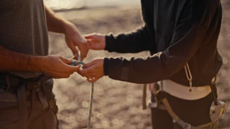 In-a-close-up-shot,-a-girl-in-a-black-gym-uniform-demonstrates-how-to-properly-tie-a-knot-with-a-thick-rope-on-a-guy's-equipment-and-belay-him-before-he-starts-climbing-a-rocky-cliff-during-a-rock-climbing-session.