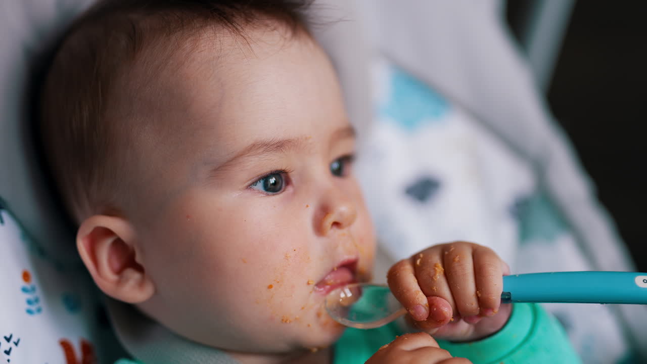 Caucasian baby dirtied with food after feeding. Little kid shoves the spoon into mouth. Close up.