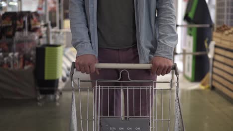 cropped front footage of a man in blue shirt and violet pants pushing the shopping empty shopping trolley the supermarket entry. the guy came for shopping to a big mall