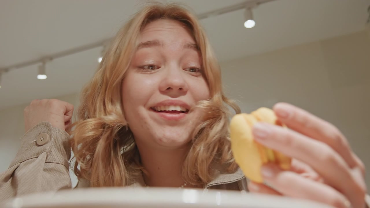 Close up of young woman with wavy blonde hair wearing beige jacket reaching for and picking up yellow macarons from plate in indoor setting. scene features soft lighting on subject face