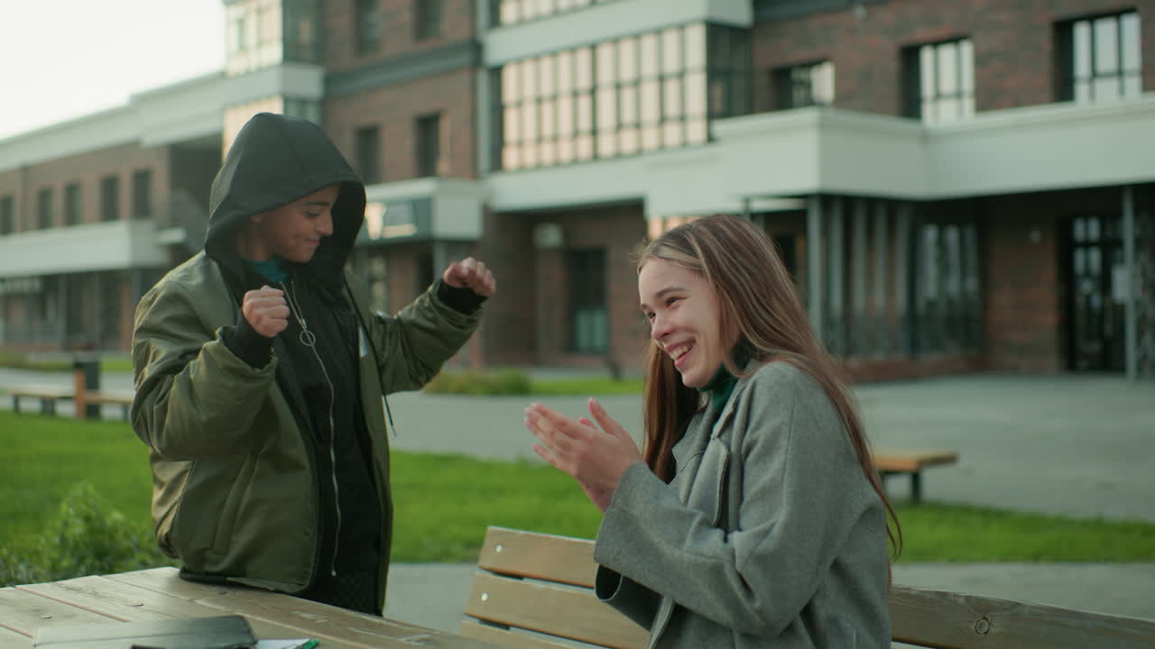 Little boy launches paper kite but it unexpectedly flies back toward his head, making his sister burst into laughter while he claps hands in playful embarrassment during outdoor fun